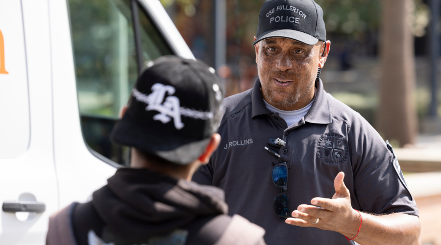 CSS Rollins talking to a student. CSS Rollins is wearing a black hat that says CSU Fullerton Police and a gray polo shirt. Only the back of the student is visible. 